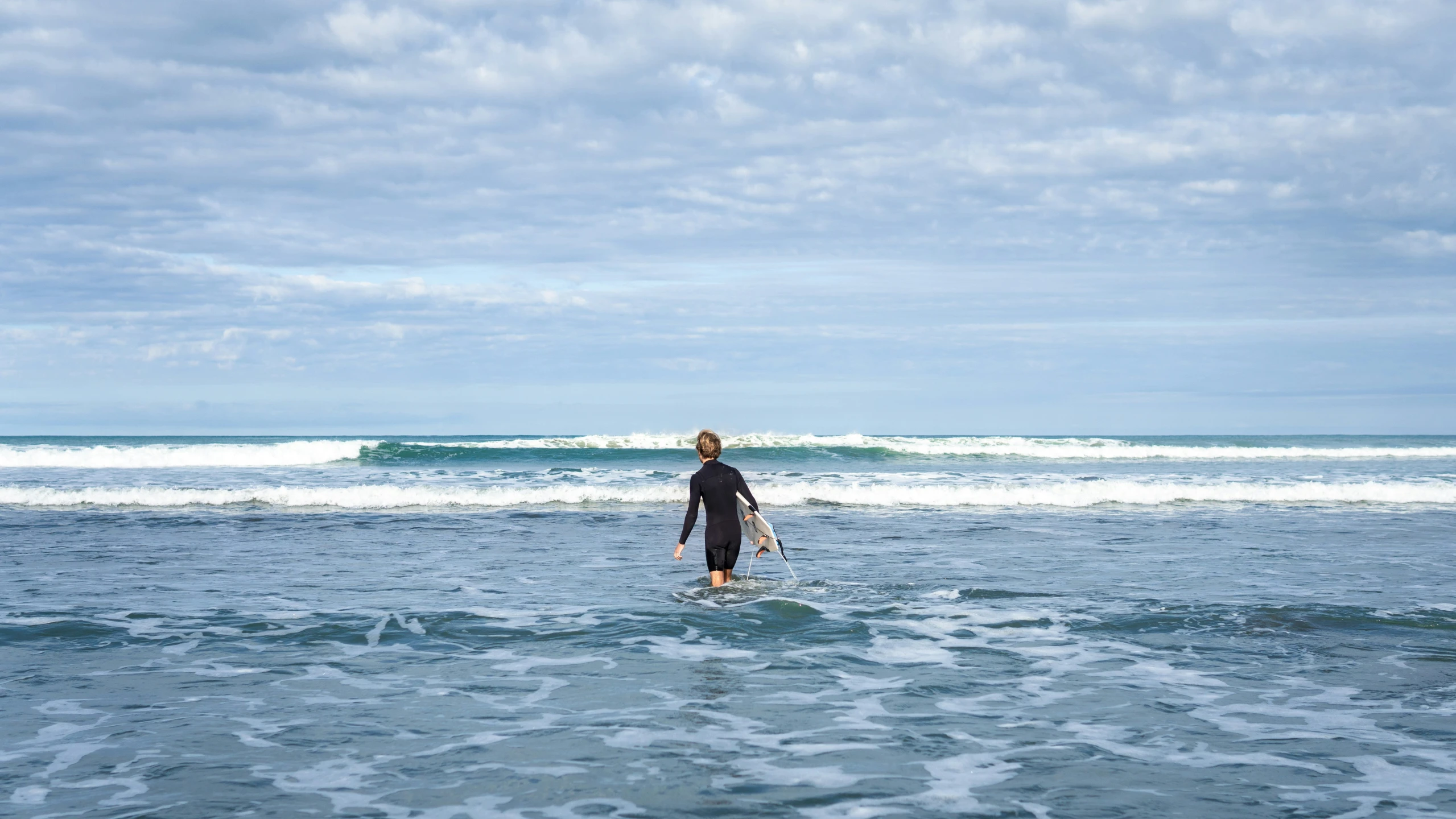 person in wetsuit wading out in the water on surfboard