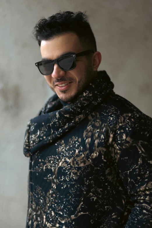 a man wearing sunglasses and jacket smiling in front of wall
