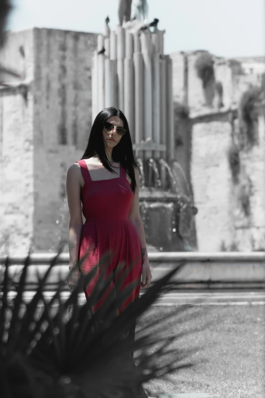 a woman with dark hair and wearing sunglasses, stands outside a fountain