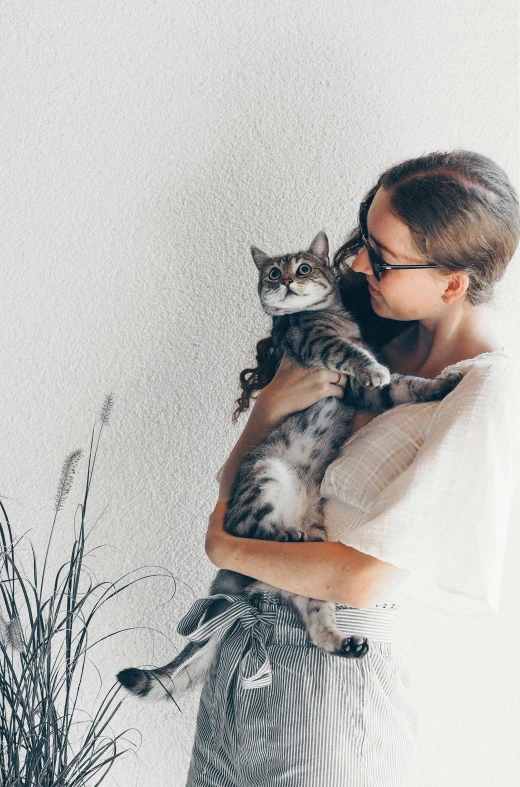 a woman wearing glasses holds a cat while standing near a plant