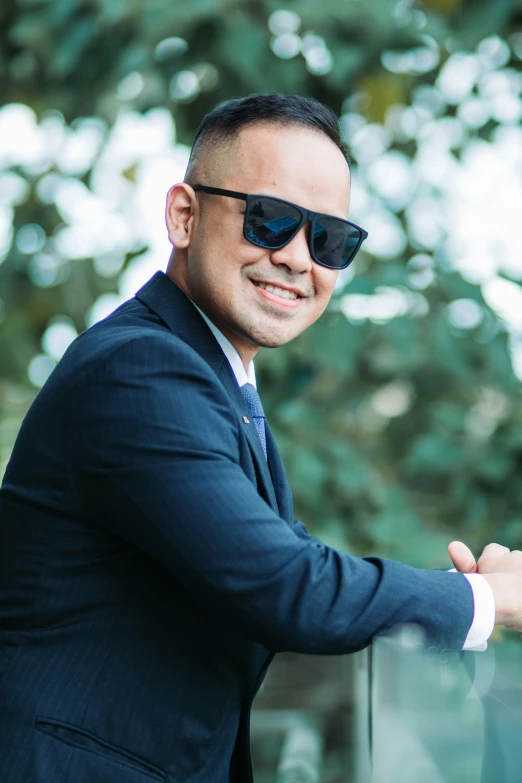 a man wearing sunglasses standing on a railing near trees