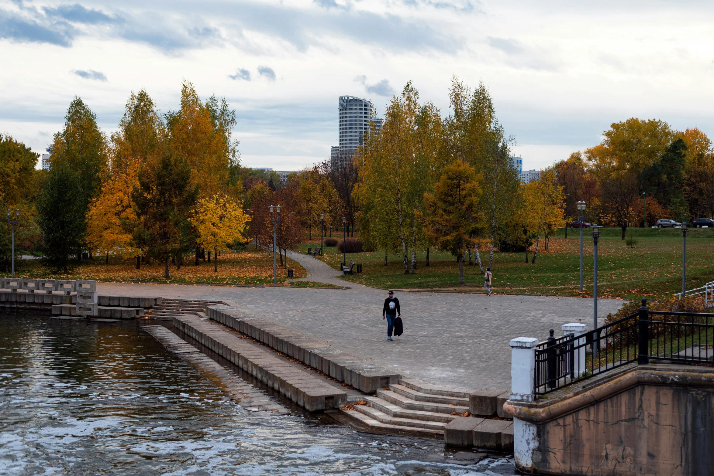 a woman in a long black dress walking across a park with some trees and a building