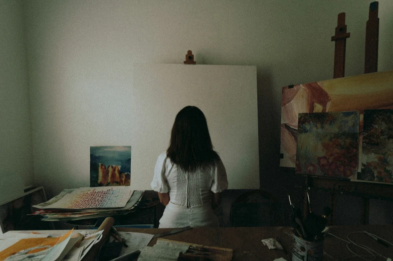 a woman with long dark hair and a white shirt stands in an art studio near some paintings