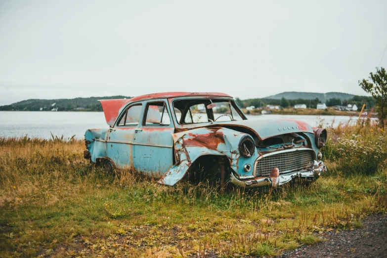 the rusty and peeling car is sitting in the grass by a body of water