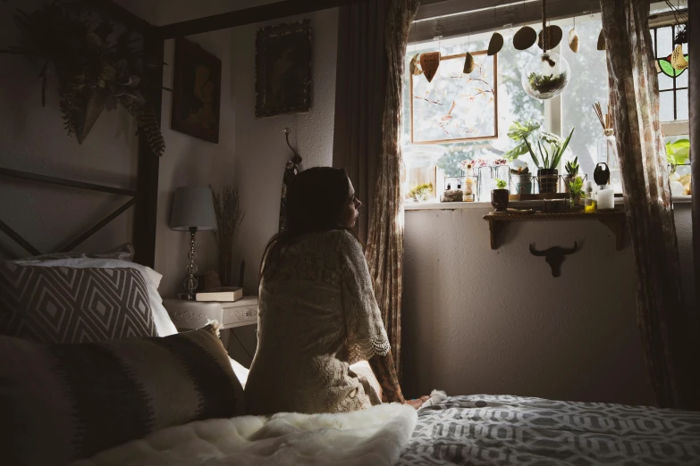 a young woman sits on the bed and looks out a window