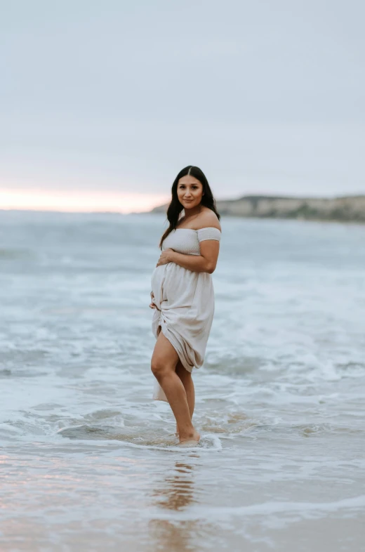 a pregnant girl stands in the shallow water at the beach