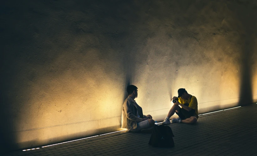 two people sitting against a wall one of them is taking his picture