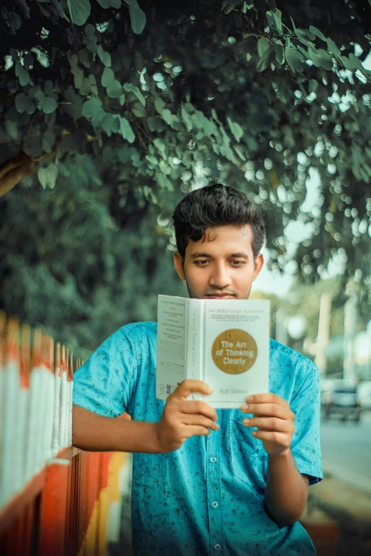 a young man stands underneath a tree holding an open book in his hands