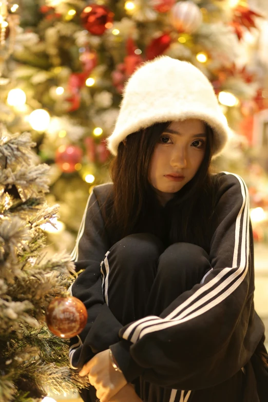 a girl wearing a winter hat sitting next to christmas tree