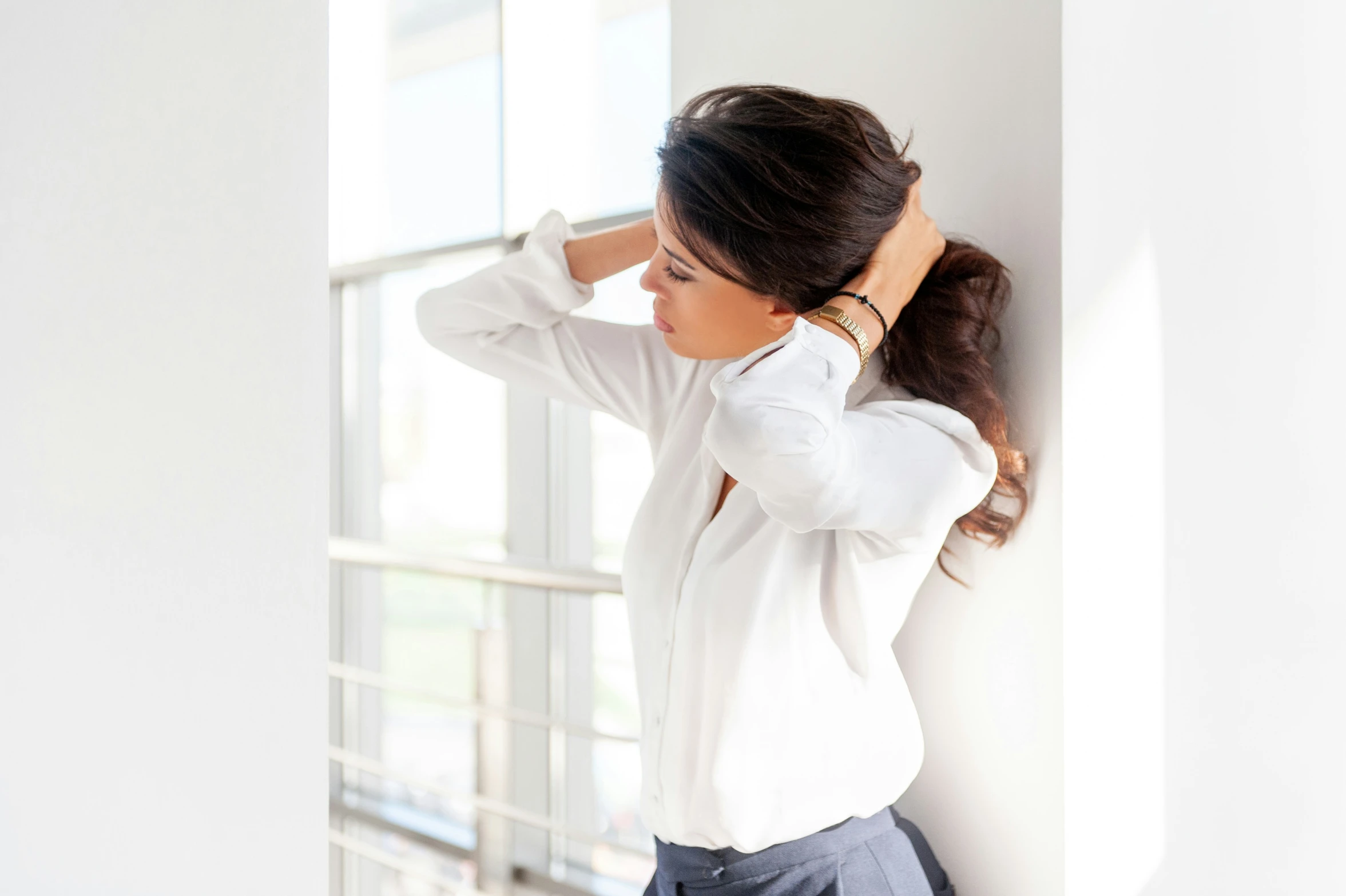 woman with long black hair leans against wall and gazes outside window