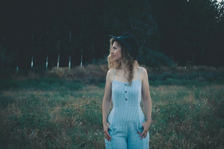 a girl is standing in a grassy field wearing overalls and a hat