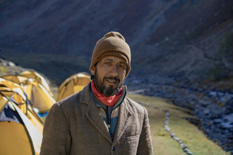 a man wearing a brown jacket and scarf and standing in front of some tents