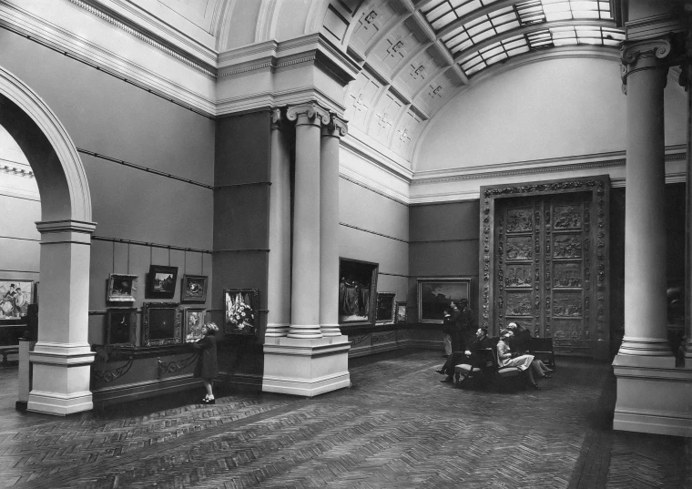 several people sitting and walking around in a room with large windows