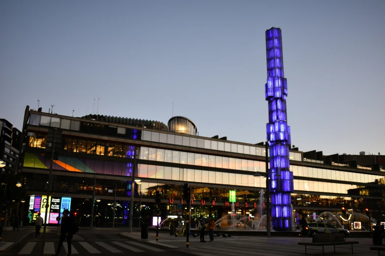 people walk near buildings and a lighted tower in the background