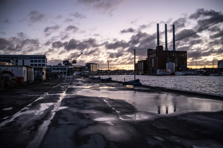 a dark looking city street with buildings, water and industrial equipment