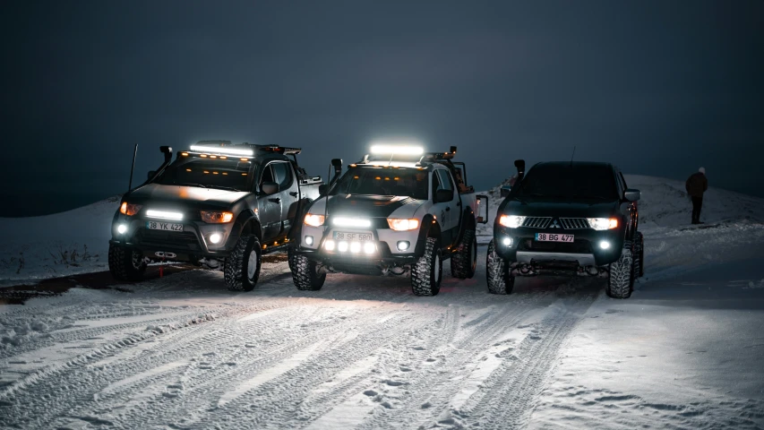 two trucks with lights on driving down a snow covered road