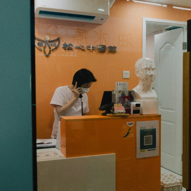 a woman with a medical mask stands behind a counter in an orange cubicle