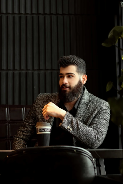 a man with beard sitting in front of a table holding a cup of coffee
