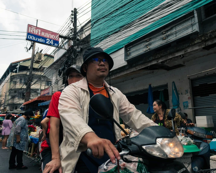 a man in an asian hat and glasses sits on his motorcycle in front of some shops