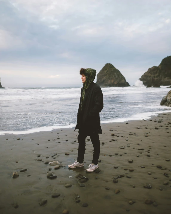 the man in black jacket stands on shore by a rocky outcropping