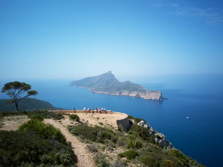 people gather on the trail along a cliff overlooking an island in the ocean