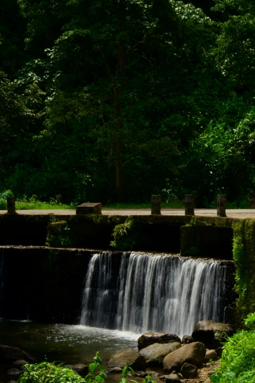 a waterfall is on one side of a road and there are trees in the background