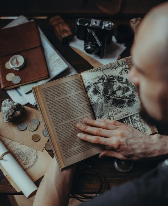 man sitting down reading a book with a camera and various craft materials