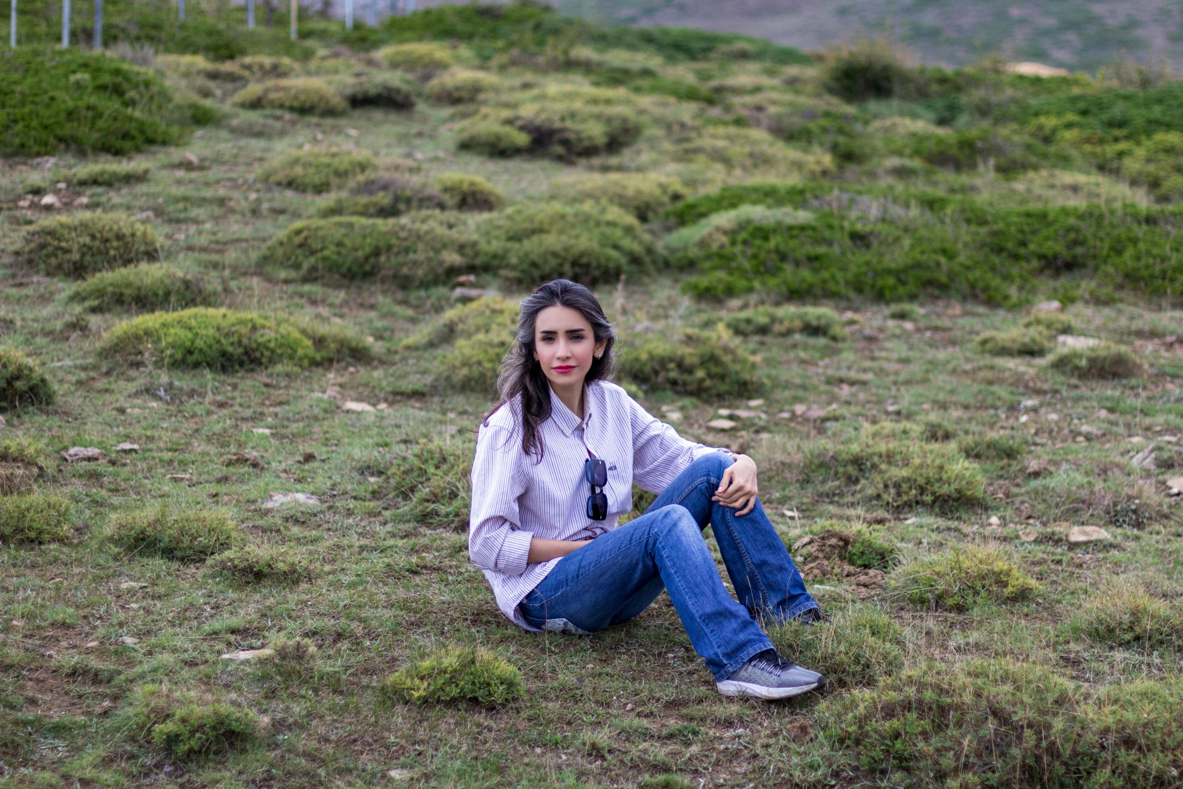 a woman in a long sleeved shirt is sitting on a grass covered hill