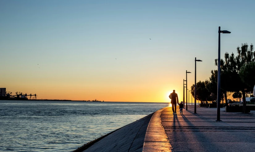 silhouette of man walking down sidewalk near water at sunset