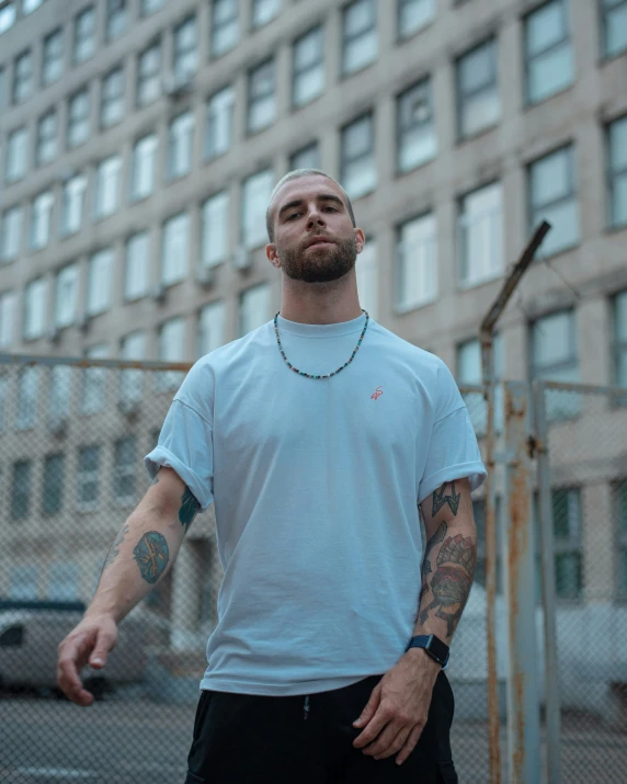 a man with tattoos stands by a fence in front of some buildings