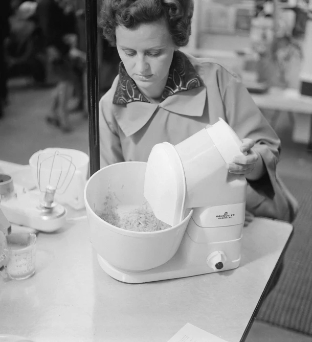 an old pograph of a woman at a kitchen counter pouring ingredients into a bowl