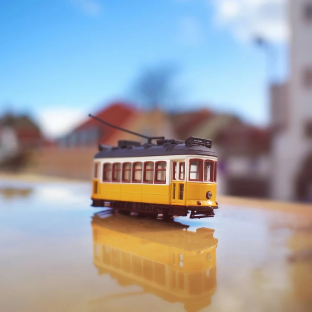 a yellow model train car sits on a surface with buildings in the background