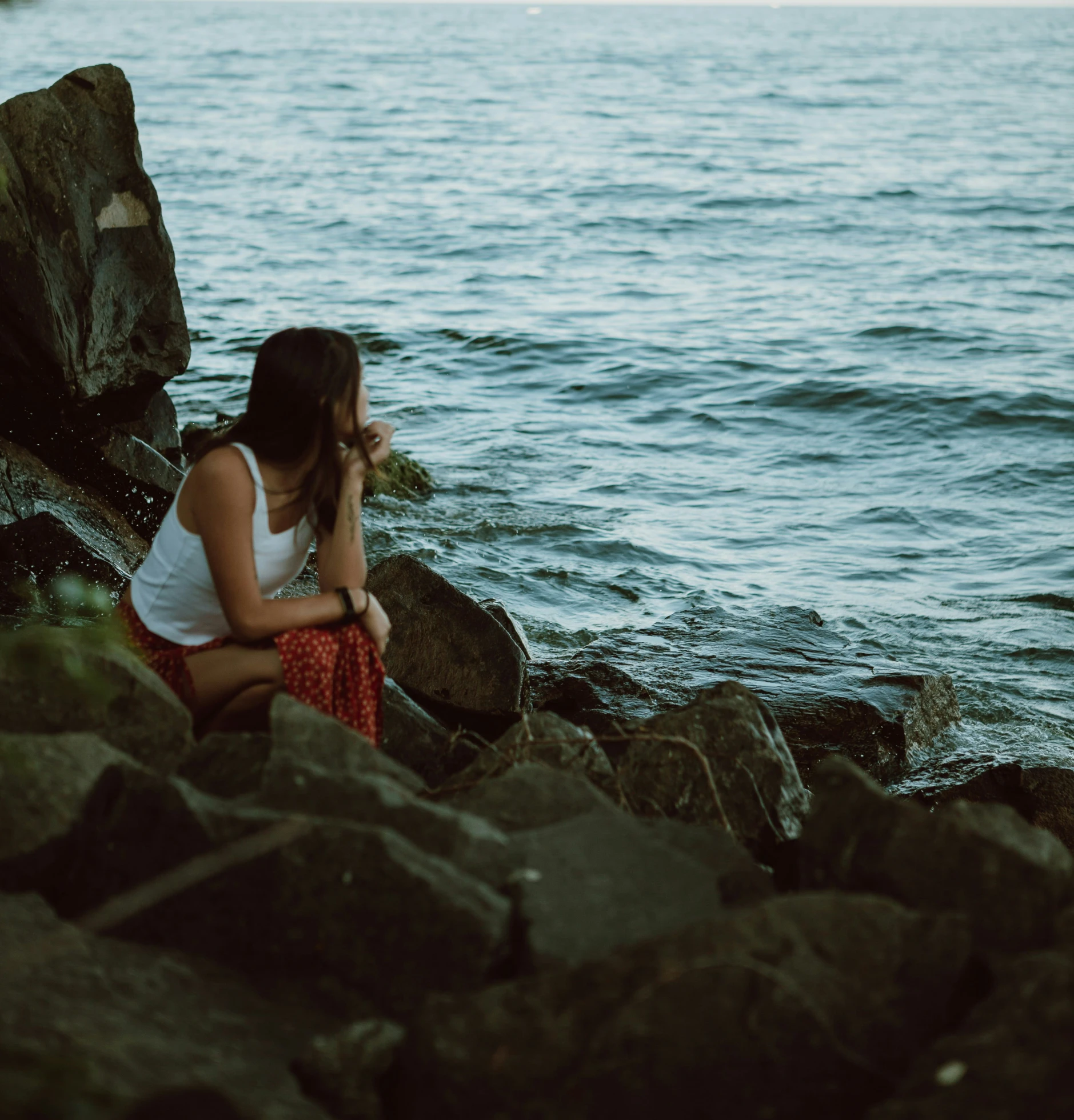 a girl sitting on a rock by the water looking at soing in her hand