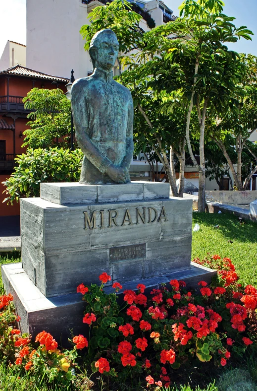 a statue in the grass near flowers with buildings in background