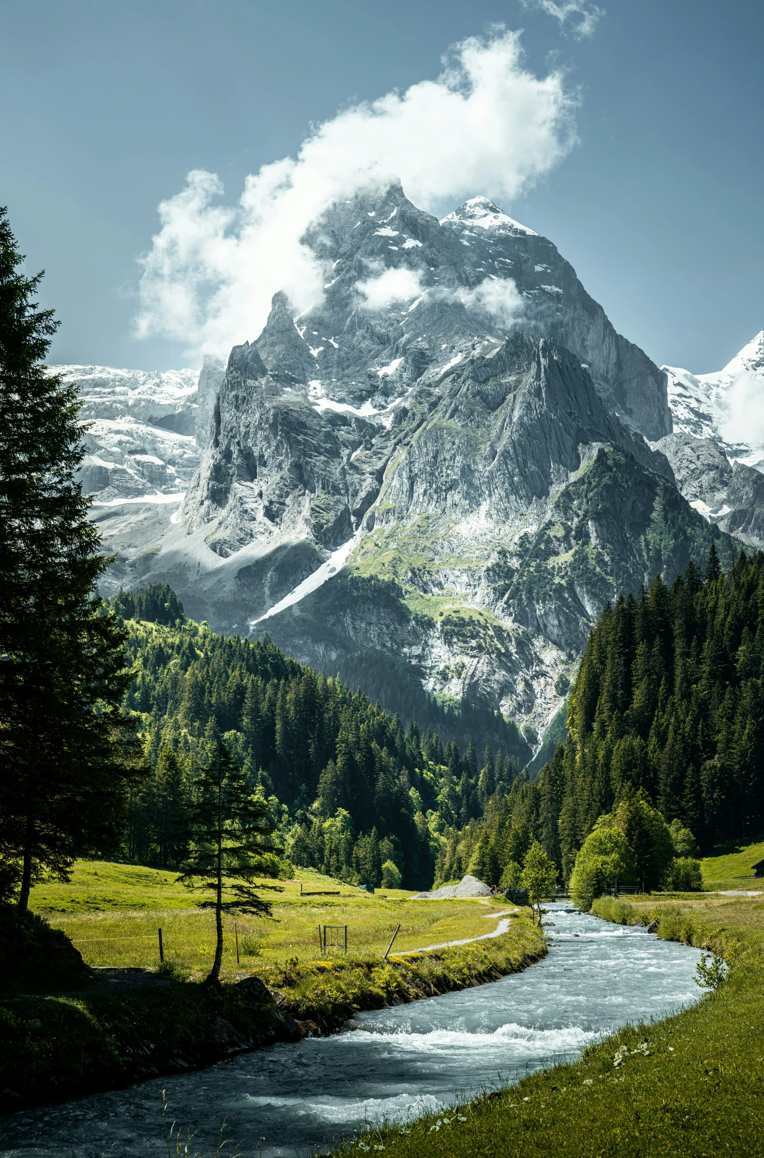 a mountain range has snow and a small stream with some green grass