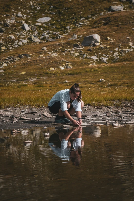 a man kneels next to a river while holding a stick