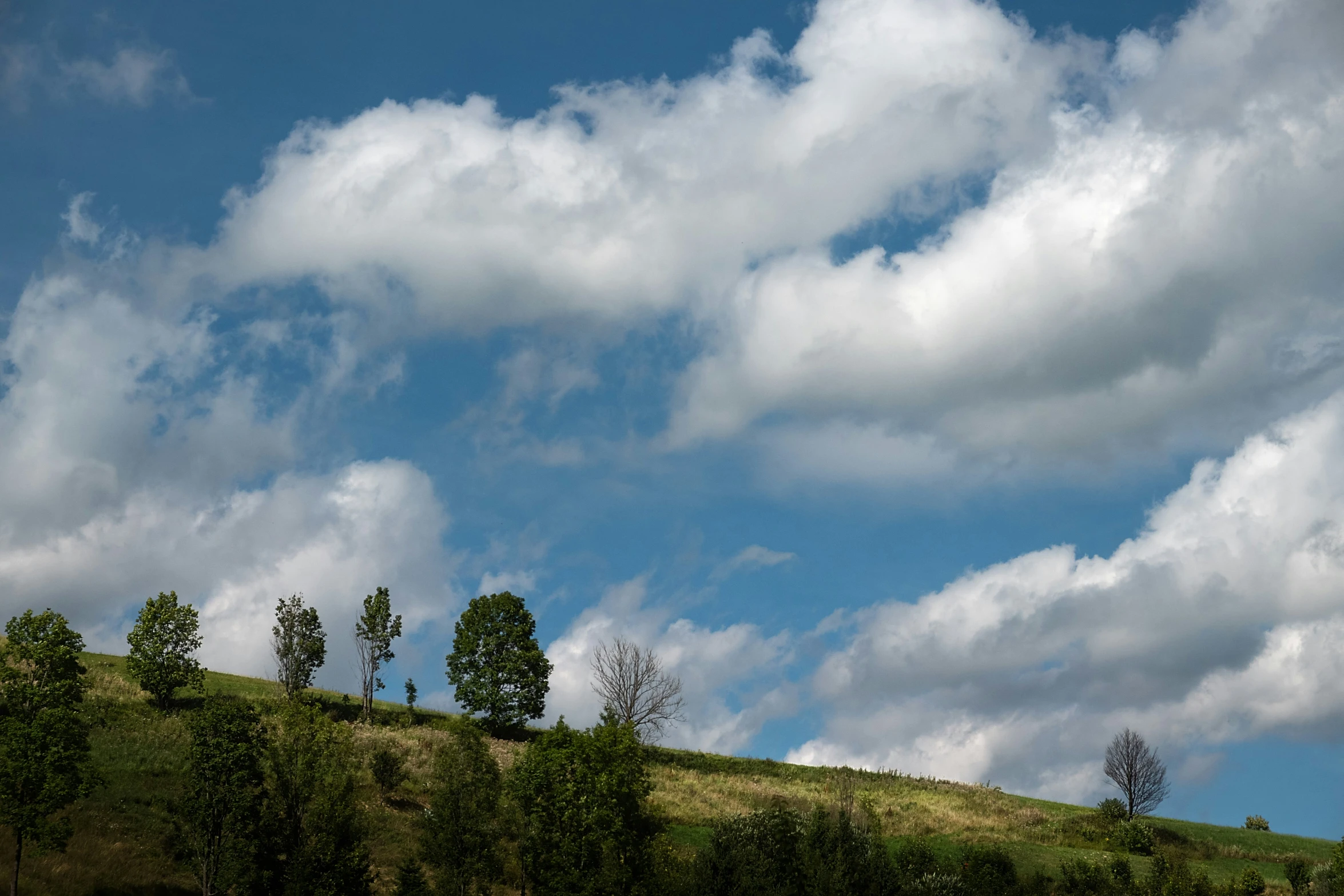 clouds rolling in to the blue sky above a hillside