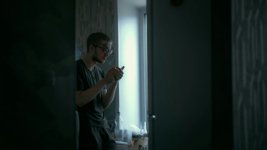 a man in glasses standing by a window while looking at his cellphone