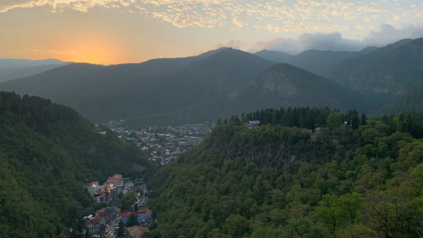 the sun sets over a city nestled between trees in the mountains