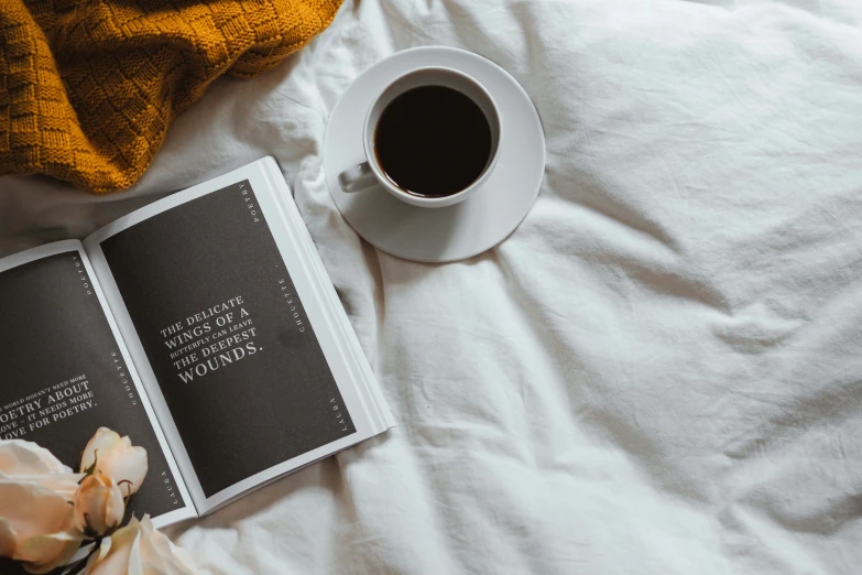 a cup of coffee sits on the bed next to an open book and flower