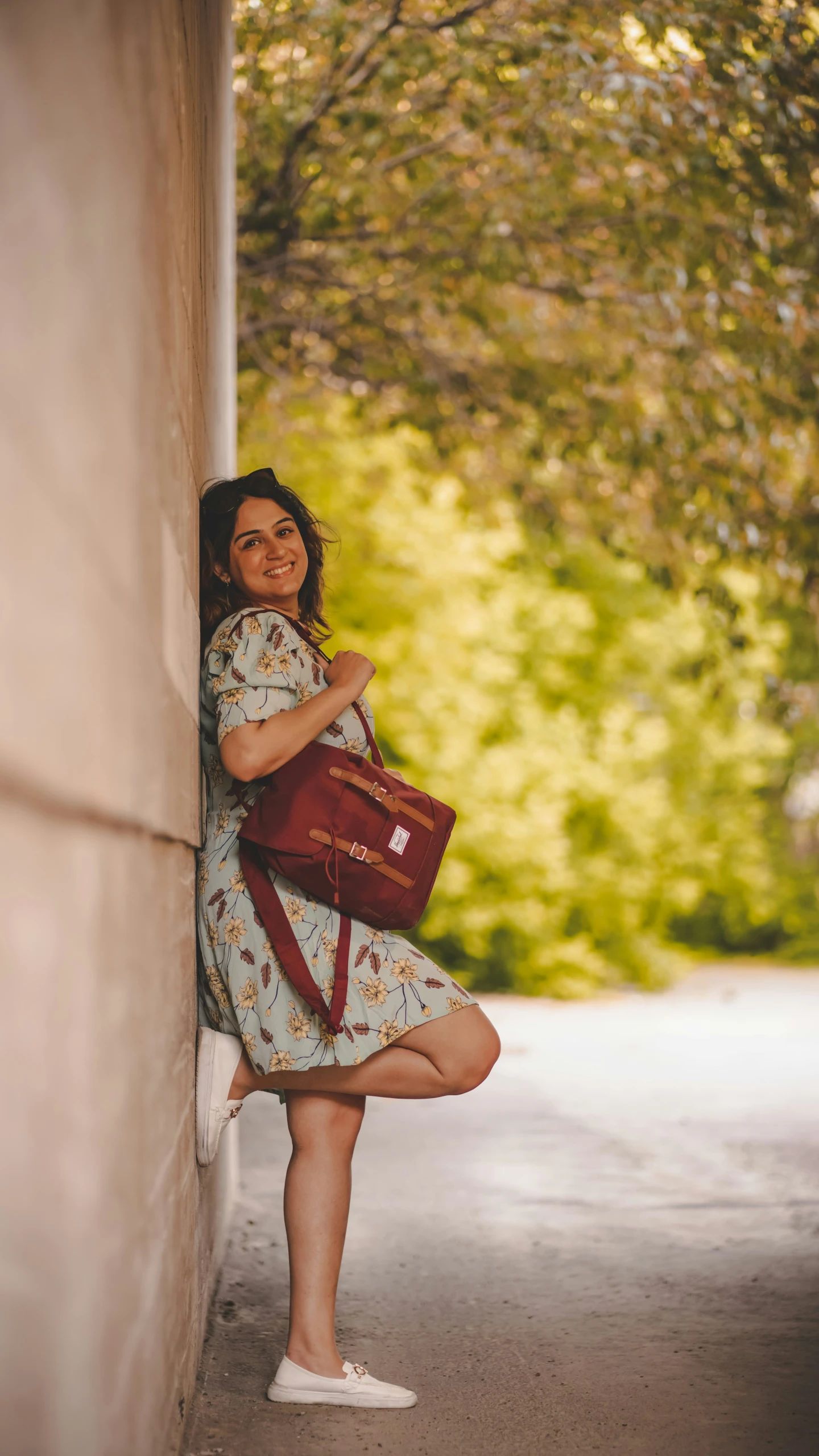 a smiling woman in dress leaning up against the wall holding a handbag