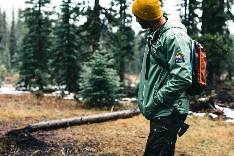 a young man in a green jacket stands in the snow near some trees