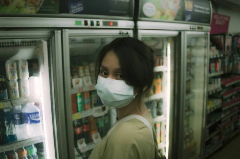 a woman in a mask stands near an upright refrigerator filled with drinks