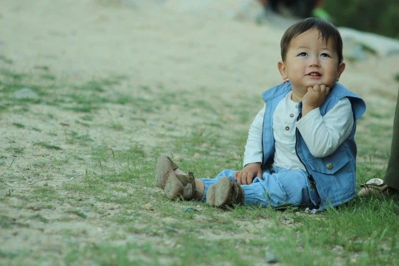 a young baby sits in a field with his hand on his mouth