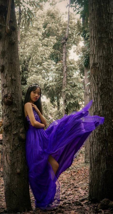 a young woman wearing a purple dress is holding her dress while standing in a forest