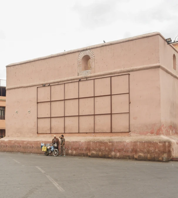 two men sitting on a motorcycle by the side of a pink building