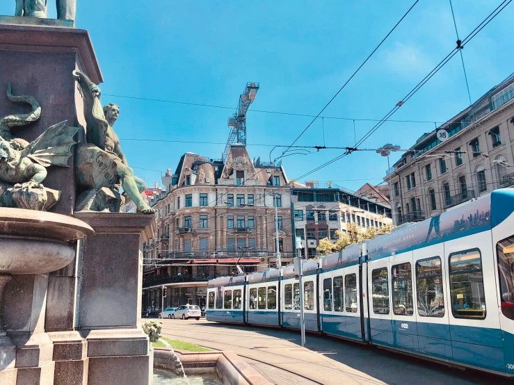 blue and white train with statue in center next to brick buildings