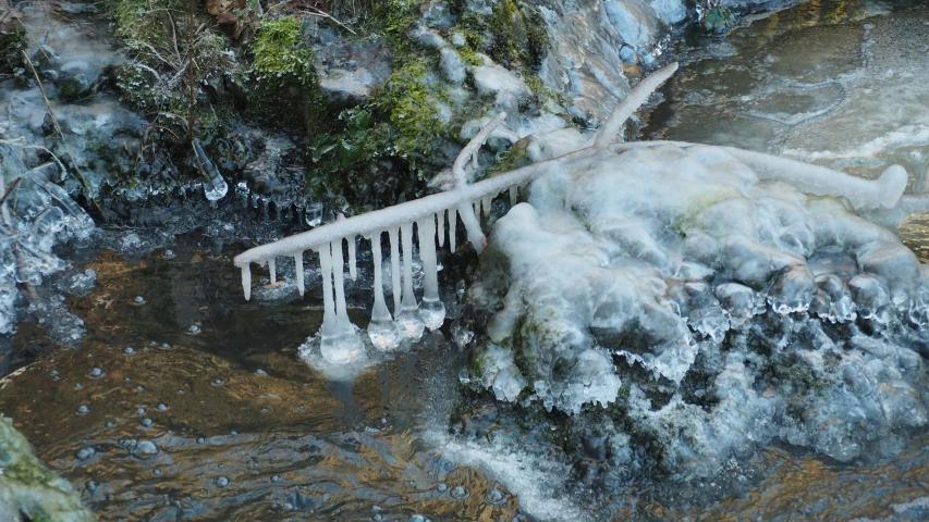 the ice - covered water has icicles on it and a stream runs along one side of it
