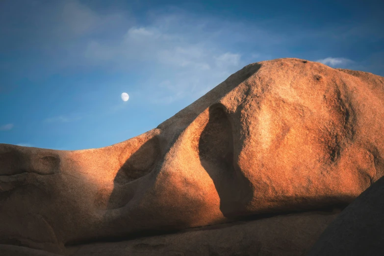 an orange rock formation with the moon in a cloudless sky