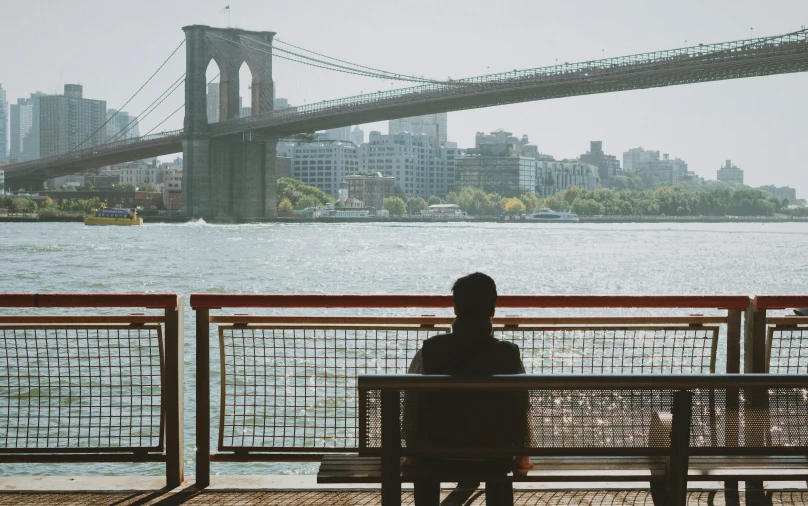 a person sits on a bench by the water in front of the bridge